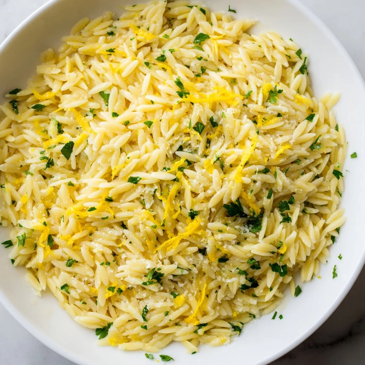 Simple lemon orzo dish topped with green parsley and parmesan shavings served in a shallow white bowl