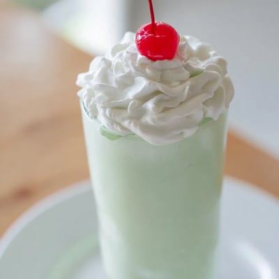 Frothy green mint milkshake blending vanilla ice cream with milk, garnished with a bright maraschino cherry for a festive American dessert.